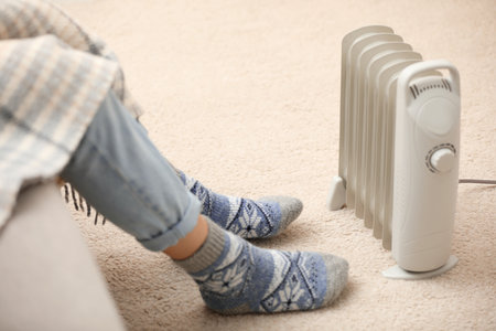 Young woman warming feet near electric heater at home, closeupの写真素材