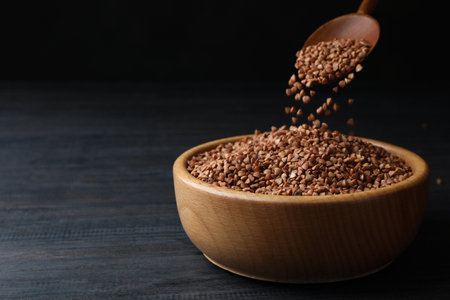 Pouring buckwheat grains from spoon into wooden bowl on gray table. Space for textの写真素材
