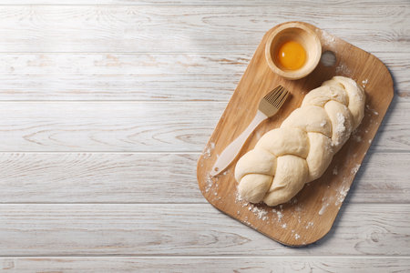 Raw braided bread, egg yolk and pastry brush on white wooden table, top view with space for text....