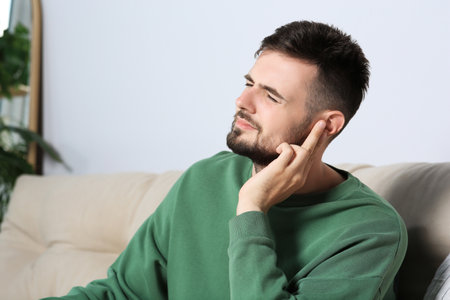 Young man suffering from ear pain at home. Space for text