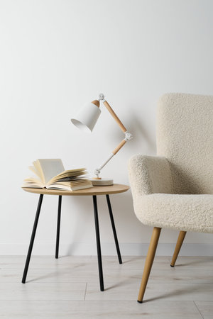 Stylish lamp with open book on wooden coffee table and soft armchair in light room. interior designの写真素材