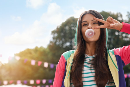 Beautiful young woman blowing chewing gum and showing peace gesture outdoors, space for textの写真素材