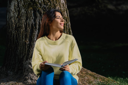 Young woman reading book near tree in park on sunny dayの写真素材