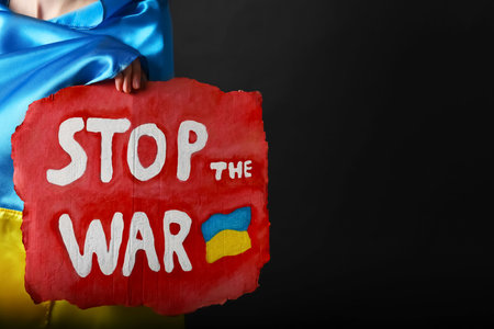 Woman wrapped in Ukrainian flag holding poster with words Stop the War on black background, closeup. Space for textの写真素材