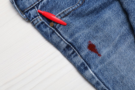 Jeans with stained red ink and pen in pocket on wooden table, top view. Space for text