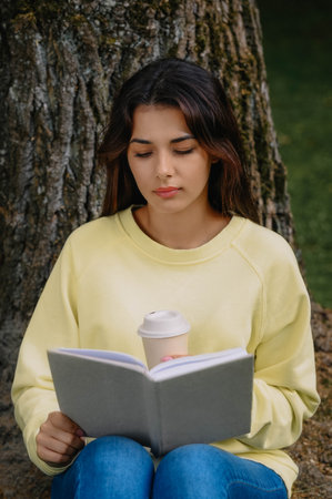 Young woman with cup of coffee reading book near tree in parkの写真素材
