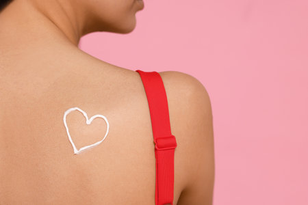 Young woman with sun protection cream on her back against pink background, closeup. Space for textの写真素材