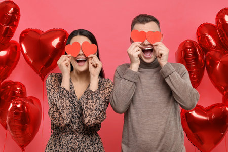 Lovely couple holding paper hearts near heart shaped air balloons on red background. Valentine's day celebrationの写真素材