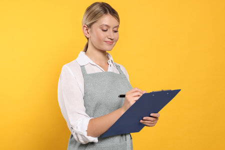 Beautiful young woman in clean apron with clipboard on orange background