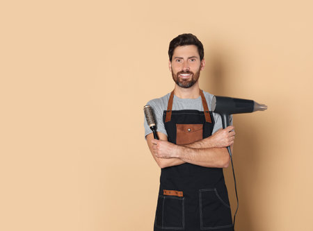 Smiling hairdresser in apron holding dryer and brush on light brown background, space for textの写真素材