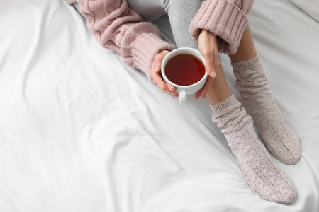 Woman with cup of tea wearing warm socks in comfortable bed, closeup. Space for textの写真素材
