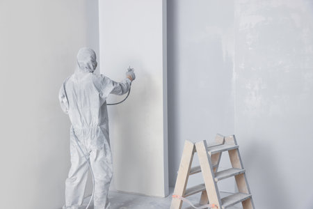 Decorator in uniform painting wall with sprayer indoors, back view