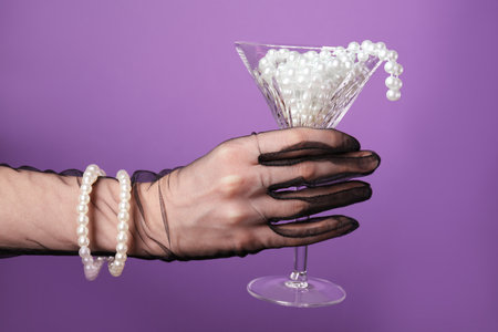 Woman in elegant gloves holding beautiful martini glass with pearls on purple background, closeupの写真素材