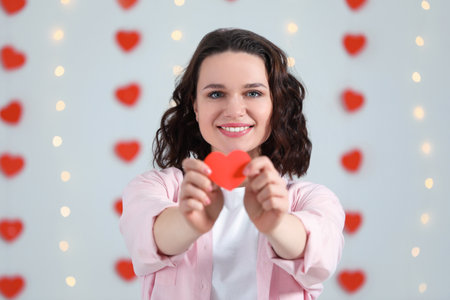 Beautiful young woman with red paper heart indoors, view from camera. Valentine's day celebration in long distance relationshipの写真素材