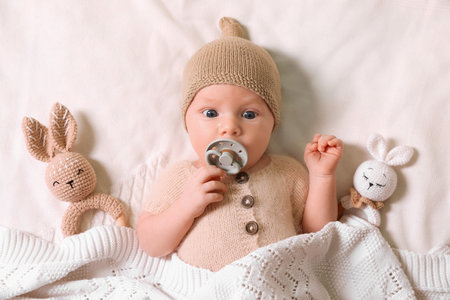 Cute newborn baby with pacifier and toys on white knitted plaid, top view