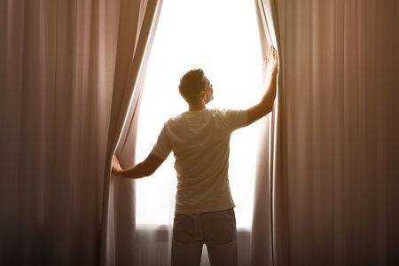 Man opening window curtains at home, back view