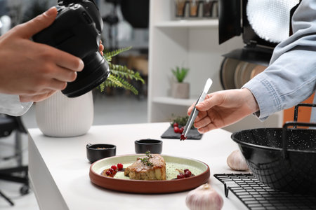 Professional photographer taking photo while food stylist decorating composition with meat medallion in studio, closeupの写真素材