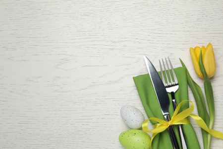 Cutlery set, painted eggs and beautiful flower on white wooden table, flat lay with space for text....