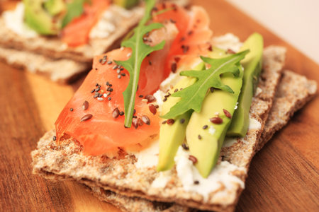 Fresh crunchy crispbreads with cream cheese, salmon, avocado and arugula on wooden board, closeupの写真素材