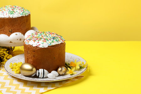 Traditional Easter cakes with sprinkles, painted eggs and beautiful spring flowers on yellow background, space for textの写真素材