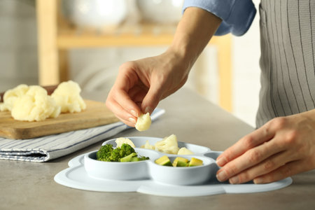 baby food. Woman putting piece of cauliflower into section plate with different vegetables at gray textured table in kitchen, closeupの写真素材