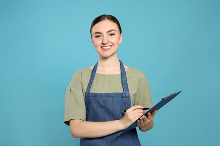 Beautiful young woman in clean denim apron with clipboard on light blue backgroundの写真素材