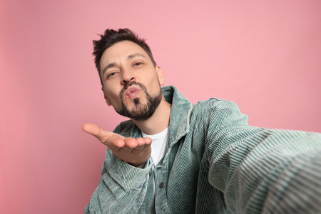 Handsome man blowing kiss while taking selfie on pink backgroundの写真素材