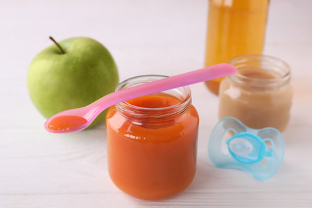 Jars with healthy baby food, bottle of juice, apple, spoon and pacifier on white wooden tableの写真素材