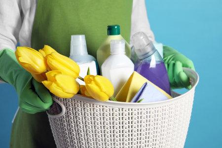 spring cleaning. Woman holding basket with detergents, flowers and rags on light blue background, closeupの写真素材