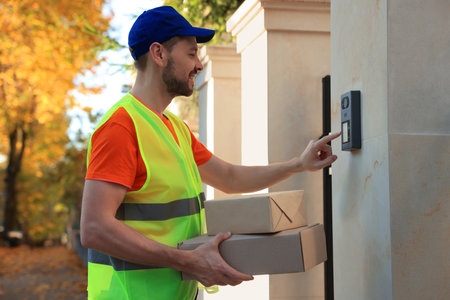 Courier in uniform with parcels ringing doorbell outdoors