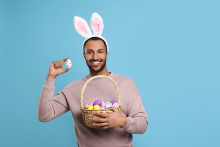 Happy African American man in bunny ears headband holding wicker basket with Easter eggs on light blue background. Space for textの写真素材
