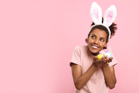 Happy African American woman in bunny ears headband holding Easter eggs on pink background, space...