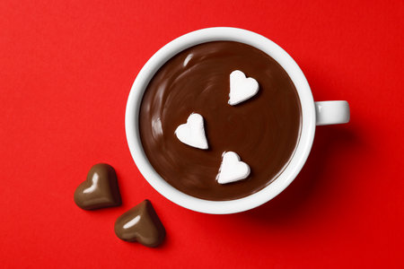 Cup of hot chocolate with heart shaped marshmallows and tasty candies on red background, flat layの写真素材