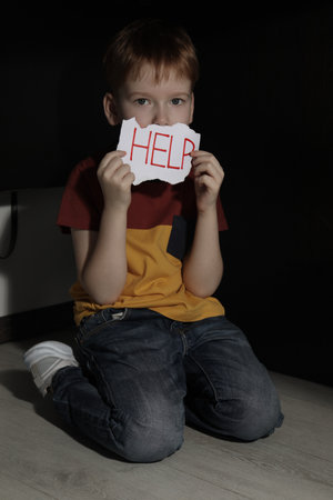 Scared little boy holding piece of paper with word Help under table on floor indoors. Domestic...