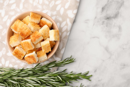 Delicious crispy croutons in bowl and rosemary on white marble table, flat lay. Space for textの写真素材