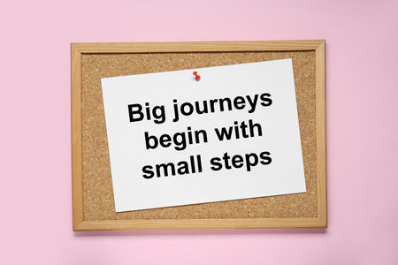 Corkboard with pinned message Big Journeys Begin With Small Steps on pink background, top view....