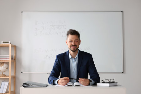 Happy teacher sitting at table in classroomの写真素材