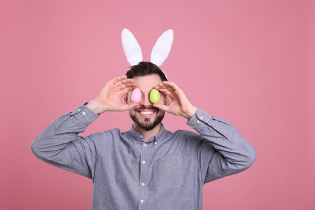 Happy man in bunny ears headband holding painted Easter eggs near his eyes on pink backgroundの写真素材