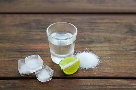 Mexican tequila shot with lime slice, ice cubes and salt on wooden table. drink made from agave
