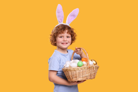 Portrait of happy boy in cute bunny ears headband holding wicker basket with Easter eggs on orange...