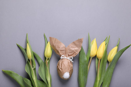 Easter bunny made of kraft paper and egg near beautiful tulips on gray background, flat lay. Space for textの写真素材