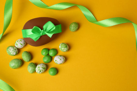 Tasty chocolate egg with green ribbon and candies on yellow background, flat lay. Space for textの写真素材