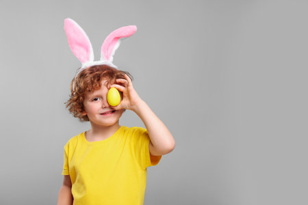 Happy boy in cute bunny ears headband covering eye with Easter egg on light grey background. Space...