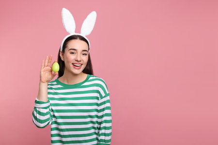 Happy woman in bunny ears headband holding painted Easter egg on pink background. Space for text.の写真素材