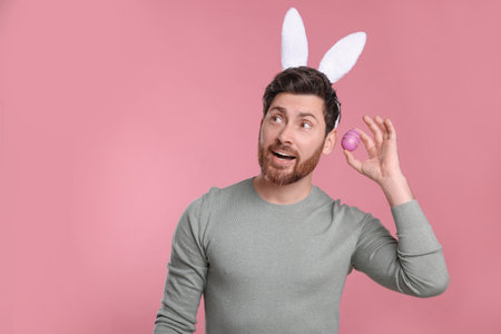 Emotional man in cute bunny ears headband holding Easter egg on pink background. Space for textの写真素材