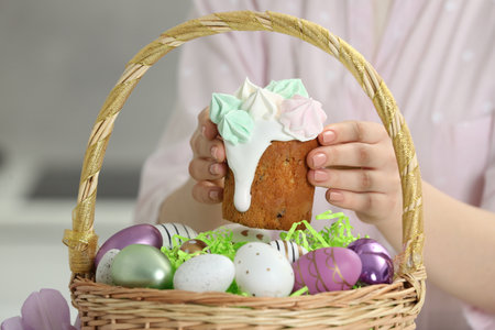 Woman putting delicious Easter cake with meringues into wicker basket with colorful eggs, closeup