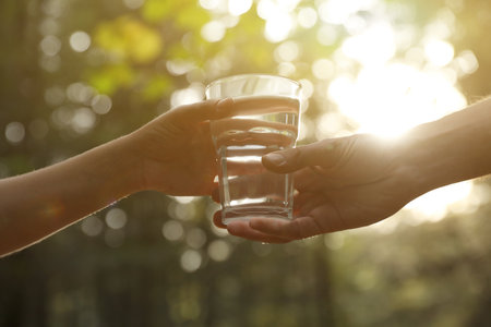 Man giving woman glass of fresh water in forest on sunny day, closeupの写真素材