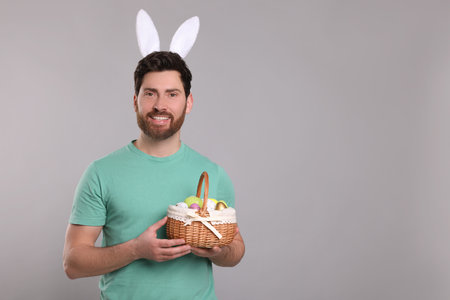 Portrait of happy man in cute bunny ears headband holding wicker basket with Easter eggs on light gray background. Space for textの写真素材