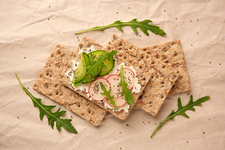 Tasty crispbreads with cream cheese, fresh cucumbers, radish and arugula on parchment paper, flat...