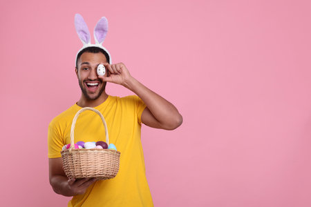 Happy African American man in bunny ears headband covering eye with Easter egg on pink background,...